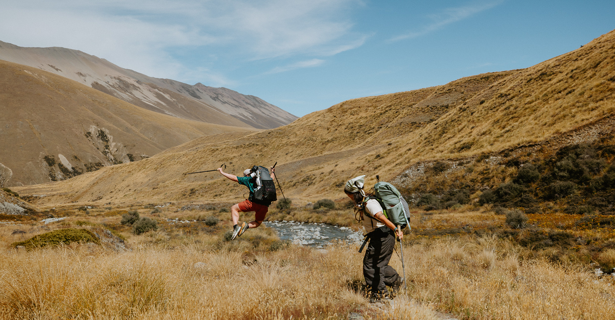 Two hikers with large backpacks traverse a grassy mountain valley under a blue sky, with one hiker jumping in the air and the other walking below.